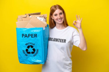Sarı arka planda izole edilmiş geri dönüşüm için elinde kağıt dolu bir geri dönüşüm çantası tutan genç kızıl saçlı kadın parmaklarıyla tamam işareti gösteriyor.