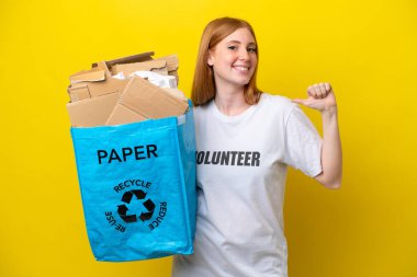 Sarı arka planda izole edilmiş, gururlu ve kendini beğenmiş bir şekilde geri dönüşüm için kağıt dolu bir çanta tutan genç kızıl saçlı kadın.