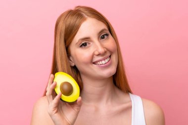 Genç kızıl saçlı kadın pembe arka planda izole edilmiş, elinde avakado tutarken gülümsüyor. Portreyi kapat