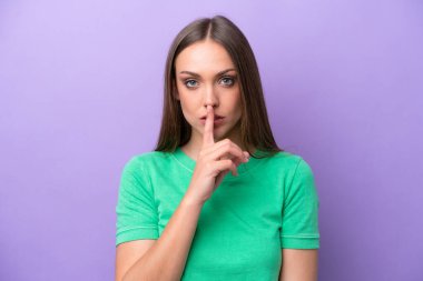 Mor arka planda izole edilmiş beyaz bir kadın sessiz bir jest işareti gösteriyor. Parmağını ağzına sokuyor.
