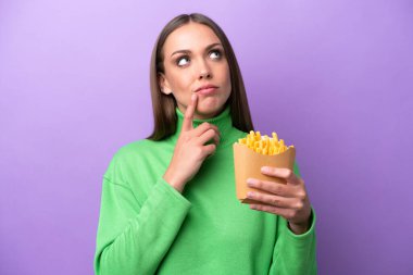 Young caucasian woman holding fried chips on purple background having doubts while looking up