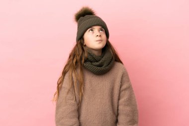 Kış şapkalı küçük kız pembe arka planda izole edilmiş ve yukarı bakıyor.