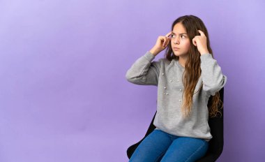 Küçük beyaz kız, mor arka planda izole edilmiş bir sandalyede oturuyor. Şüpheleri var ve düşünüyor.