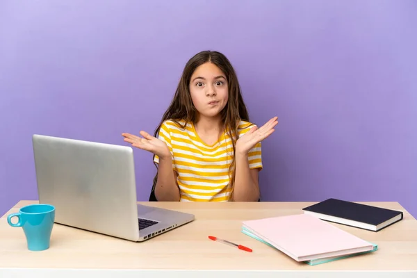 Little student girl in a workplace with a laptop isolated on purple background having doubts while raising hands