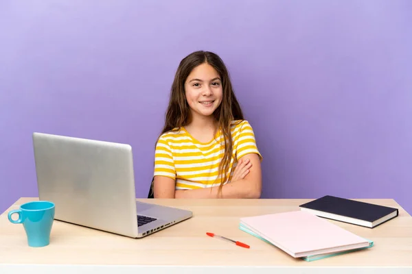 Little student girl in a workplace with a laptop isolated on purple background keeping the arms crossed in frontal position