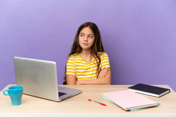 Little student girl in a workplace with a laptop isolated on purple background looking to the side
