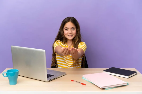 Little student girl in a workplace with a laptop isolated on purple background holding copyspace imaginary on the palm to insert an ad