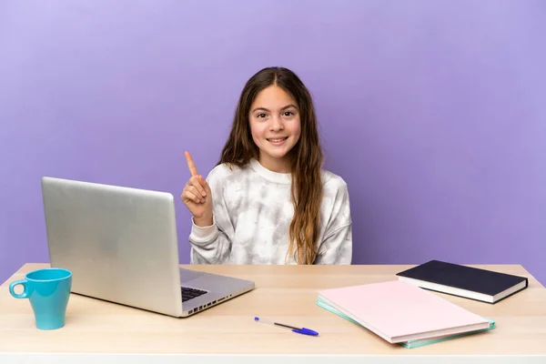 Little student girl in a workplace with a laptop isolated on purple background pointing up a great idea