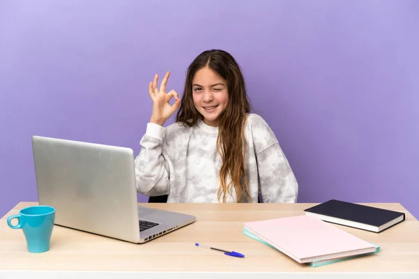 Little student girl in a workplace with a laptop isolated on purple background showing ok sign with fingers