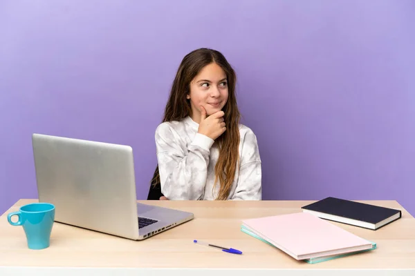 Little student girl in a workplace with a laptop isolated on purple background thinking an idea while looking up