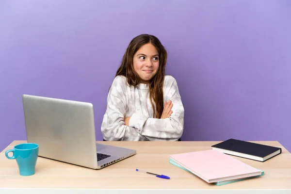 Little student girl in a workplace with a laptop isolated on purple background making doubts gesture while lifting the shoulders