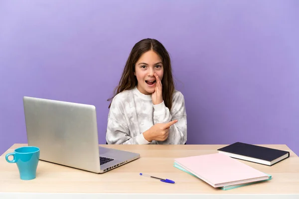 Little student girl in a workplace with a laptop isolated on purple background pointing to the side to present a product and whispering something