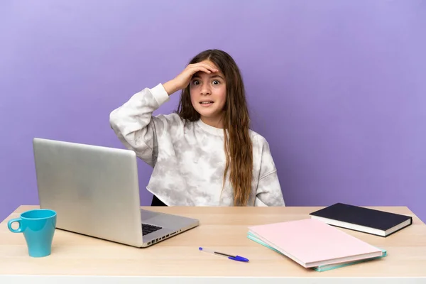 Little student girl in a workplace with a laptop isolated on purple background doing surprise gesture while looking front