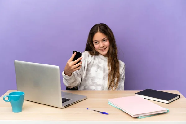 Little student girl in a workplace with a laptop isolated on purple background making a selfie