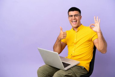 Genç adam dizüstü bilgisayarlı bir sandalyede oturuyor. Tamam işareti yapıyor ve başparmağını kaldırıyor.