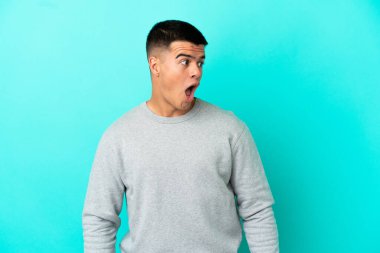 Soyutlanmış mavi arka planda genç yakışıklı bir adam yan tarafa bakarken sürpriz jest yapıyor.