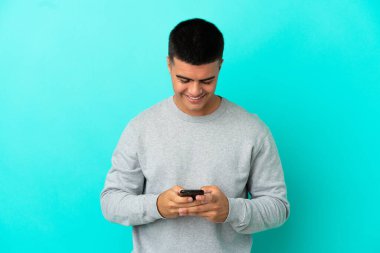 İzole edilmiş mavi arka planda genç yakışıklı bir adam cep telefonuyla bir mesaj gönderiyor.