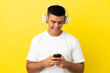 Young handsome man over isolated yellow background listening music and looking to mobile