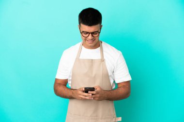 Restaurant waiter man over isolated blue background sending a message with the mobile