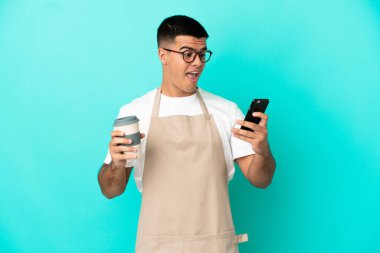 Restoran garsonu mavi arka planda. Elinde kahve ve cep telefonu var.