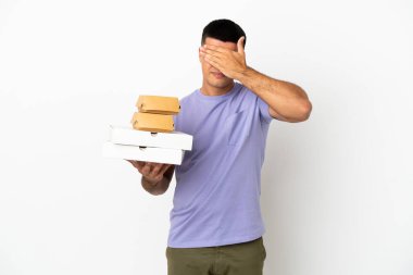 Young handsome man holding pizzas and burgers over isolated white background covering eyes by hands. Do not want to see something