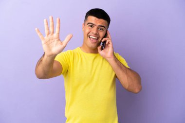 Young handsome man using mobile phone over isolated purple background counting five with fingers