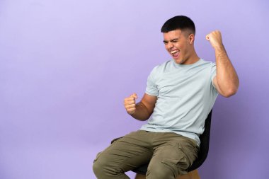 Young man sitting on a chair over isolated background celebrating a victory