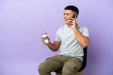 Arka planda izole edilmiş bir sandalyede oturan genç bir adam kahve ve bir cep telefonu tutuyor.