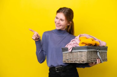 Young English woman holding a clothes basket isolated on yellow background pointing finger to the side and presenting a product