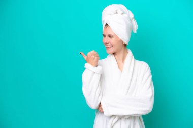 Mavi arka planda izole edilmiş bornozlu genç bir kadın bir ürünü sunmak için kenarı işaret ediyor.