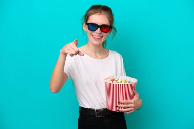 Genç İngiliz kadın mavi arka planda gözlüklü ve elinde patlamış mısırla ön tarafı işaret ediyor.