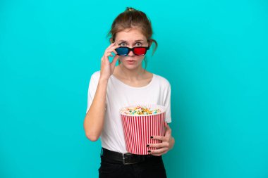 Mavi arka planda izole edilmiş genç bir İngiliz kadın 3D gözlüklü ve elinde koca bir kova patlamış mısırla şaşırdı.