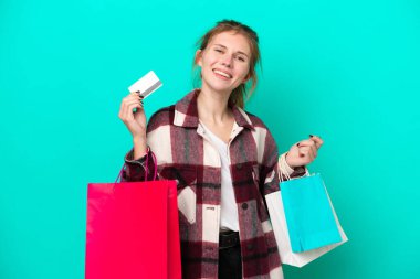 Mavi arka planda izole edilmiş genç bir İngiliz kadın elinde alışveriş çantaları ve bir kredi kartı tutuyor.