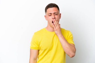 Genç Brezilyalı bir adam beyaz arka planda izole edilmiş esniyor ve geniş ağzını eliyle kapatıyor.