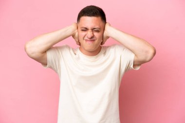 Pembe arka planda izole edilmiş genç Brezilyalı adam hayal kırıklığına uğramış ve kulaklarını kapatmış.
