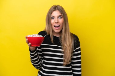 Young caucasian woman holding a bowl of cereals isolated on yellow background with surprise and shocked facial expression