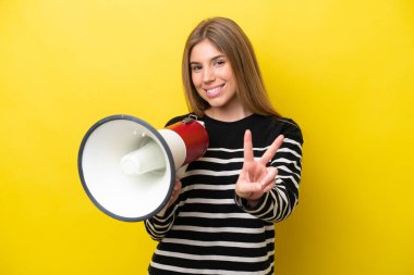 Young caucasian woman isolated on yellow background holding a megaphone and smiling and showing victory sign