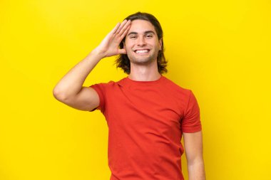 Beyaz tenli yakışıklı, sarı arka planda izole edilmiş mutlu bir ifadeyle selam veriyor.