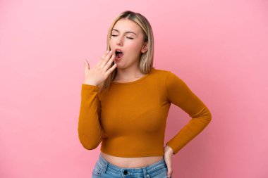 Pembe arka planda izole edilmiş genç beyaz bir kadın esniyor ve geniş bir ağzı eliyle kapatıyor.