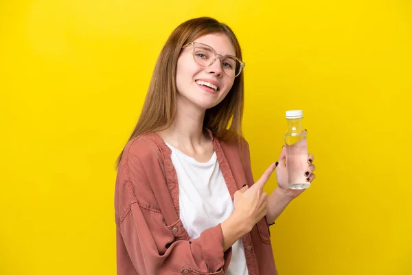 Young English woman with a bottle of water isolated on yellow background pointing back