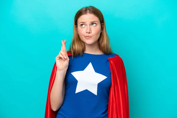 Super Hero caucasian woman isolated on blue background with fingers crossing and wishing the best