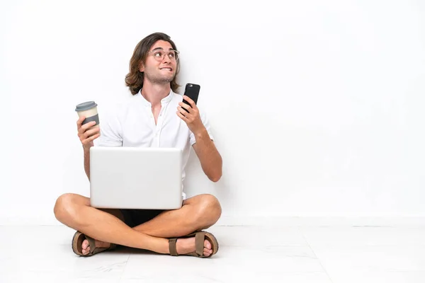 Young handsome man with a laptop sitting on the floor isolated on white background holding coffee to take away and a mobile while thinking something