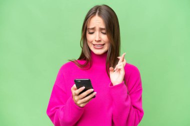 Young beautiful caucasian woman over isolated background using mobile phone with fingers crossing