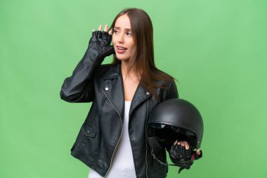 Young pretty caucasian woman with a motorcycle helmet over isolated background listening to something by putting hand on the ear
