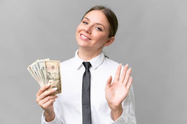 Young business caucasian woman holding money over isolated background saluting with hand with happy expression