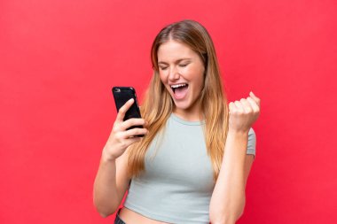 Young beautiful woman isolated on red background with phone in victory position