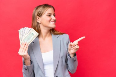 Young business caucasian woman holding money isolated on red background pointing finger to the side and presenting a product