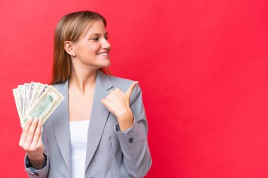 Young business caucasian woman holding money isolated on red background pointing to the side to present a product