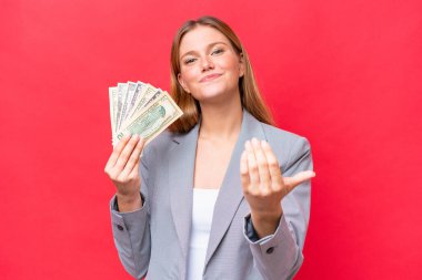 Young business caucasian woman holding money isolated on red background inviting to come with hand. Happy that you came