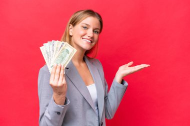 Kafkasyalı genç bir kadın kırmızı arka planda izole edilmiş parayı tutuyor ve davet etmek için ellerini yana uzatıyor.